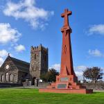 The finished memorial in weathered terracotta render, with Andreas church behind it under a blue sky