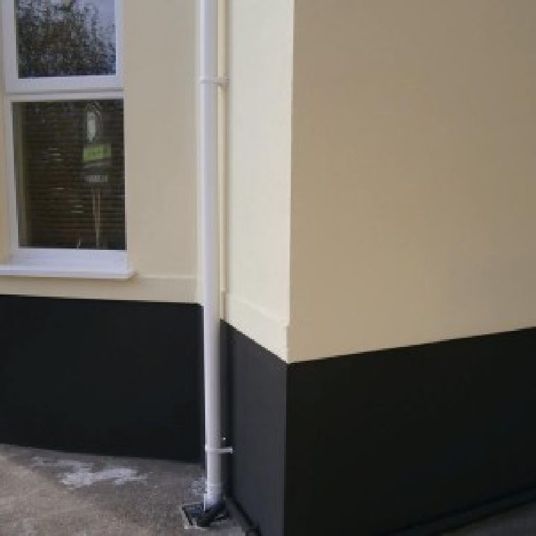 Cream render meeting the dark-painted plinth at a corner with a new downpipe