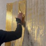 A sheet of gold foil pressed by hand against the textured panel, half of it already transferred