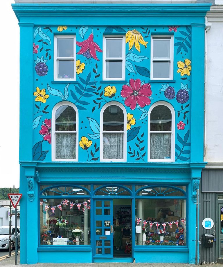The front elevation head-on, flowers across the upper floors and the shopfront in turquoise