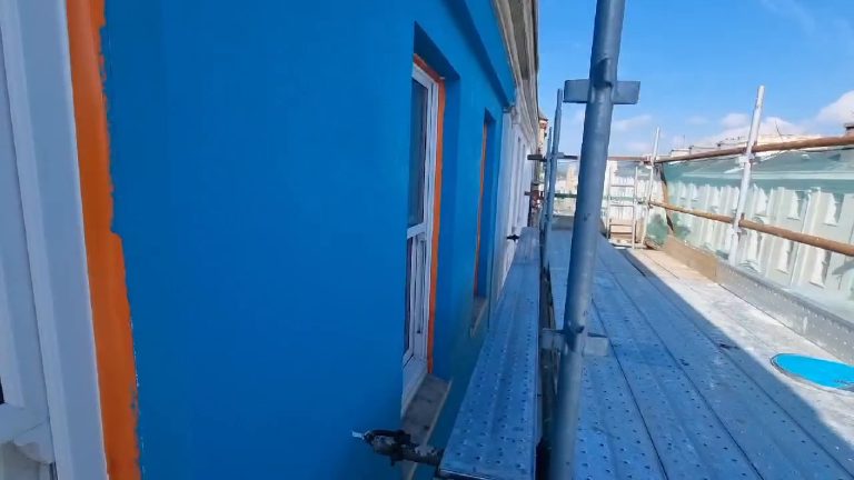 Flat turquoise render along the upper storey, windows masked in orange tape, scaffold running past